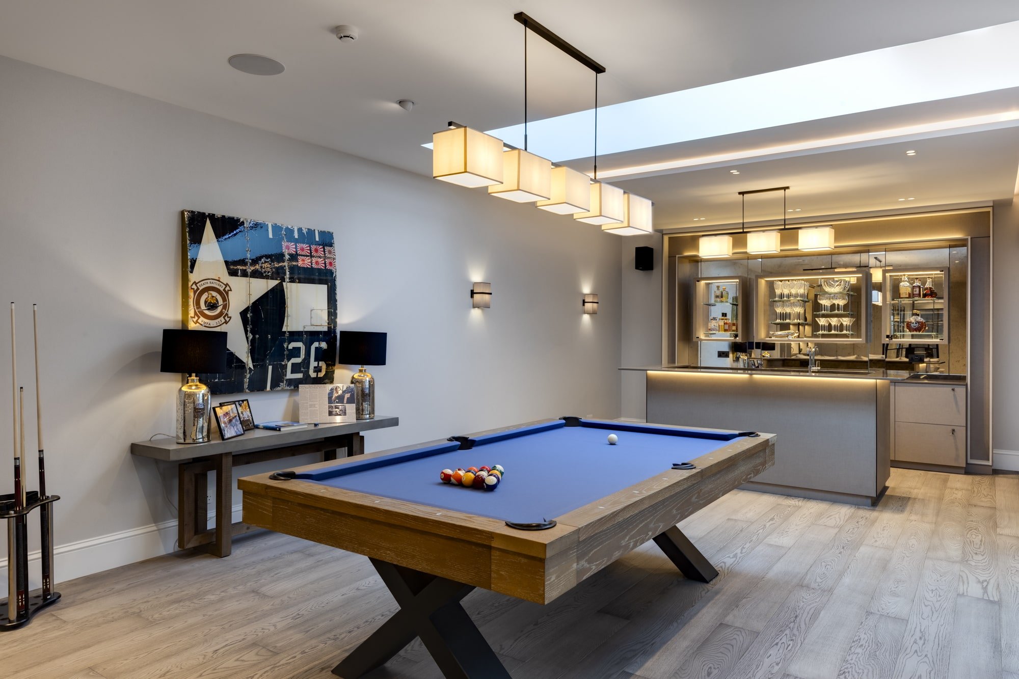 An modern basement extension with a pool table in the middle of the room infront of a mini bar next to a side table displaying lighting and a wall graphic