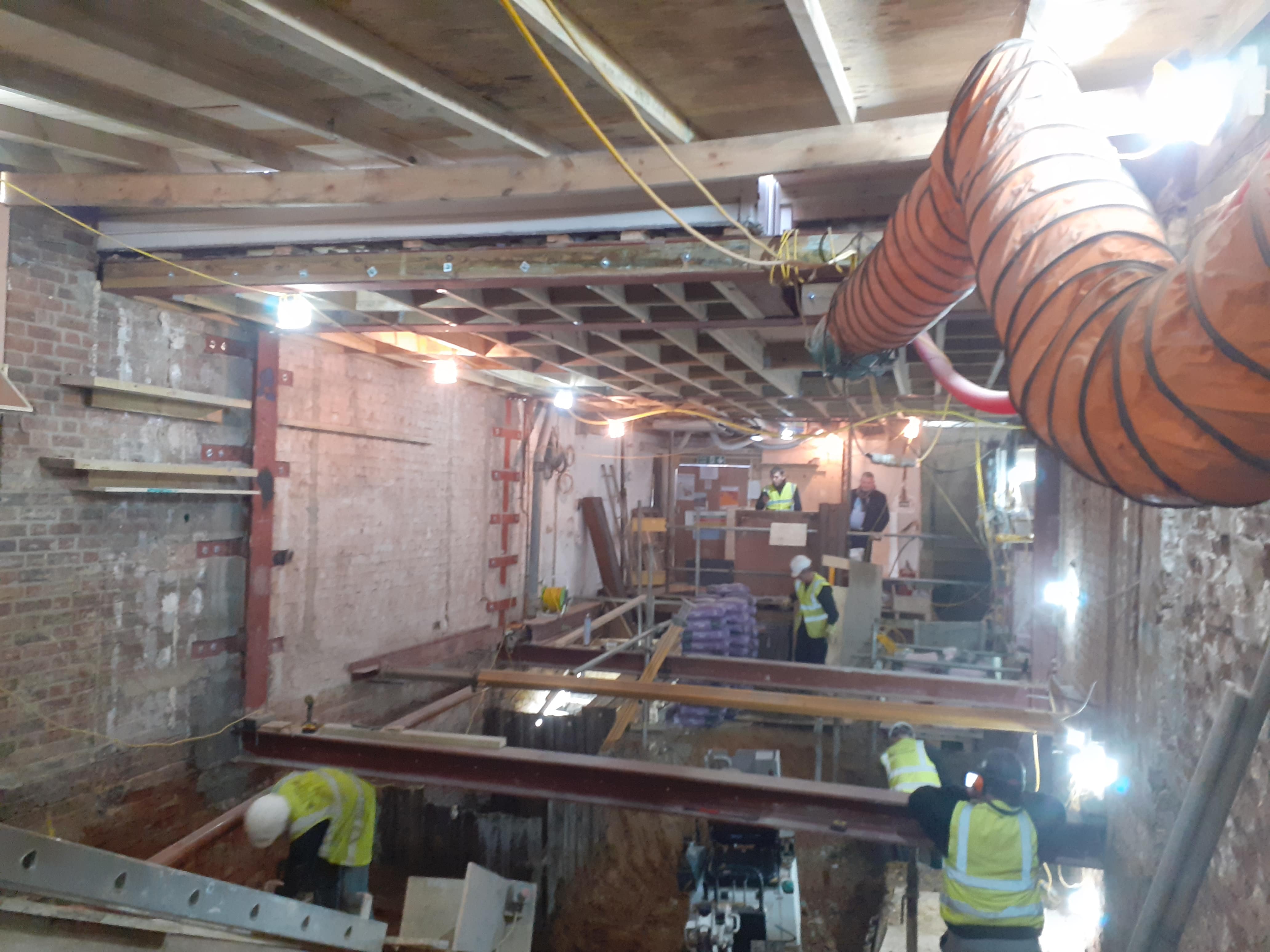 A construction site showing a basement being converted into an extension with men in high visibility jackets working on metal and wooden beams