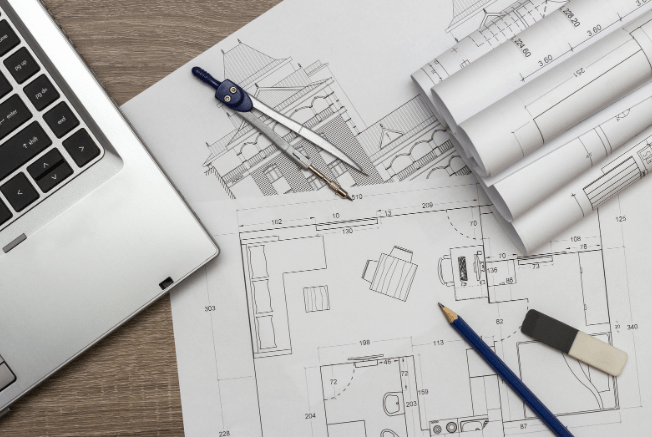 Blueprints spread out beside a laptop with a pencil and drafting compass on top highlighting the processes of modern architectural planning tools.